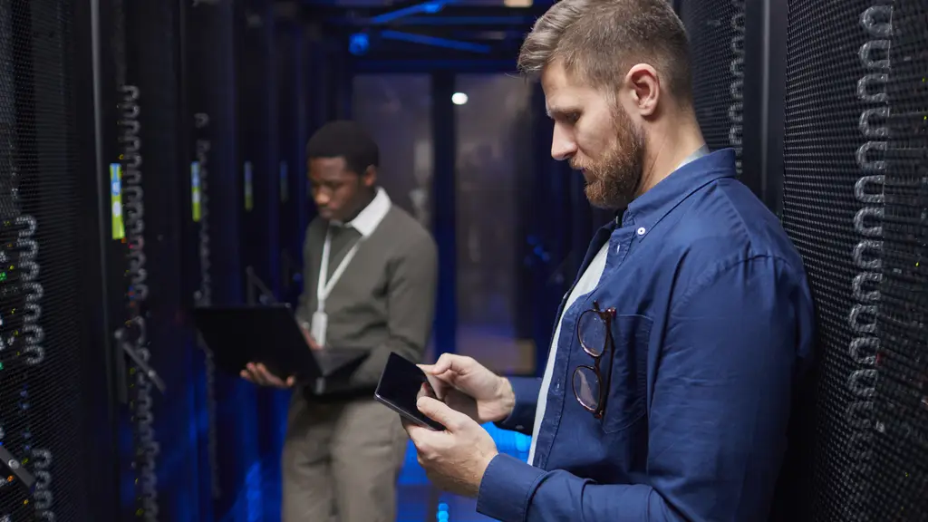 Two engineers in a server room, one with a laptop and one with a tablet